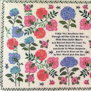 May include: Cross-stitch embroidery featuring a floral design with pink, red, and blue flowers, green leaves, and the text of a prayer. The flowers are arranged around the text, with a decorative border.