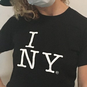May include: A person wearing a black t-shirt with the text "I NY" in white. The person is wearing a green hat and a blue face mask.