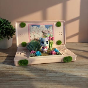 Op de afbeelding: Een lichtroze Nintendo DS-console, omgebouwd tot een miniatuurtuin. Het scherm toont een bloemrijke scène. De tuin heeft een klein karakterfiguur, vetplanten, roze en blauwe bloemen en groene mosaccenten. De console staat op een houten oppervlak.