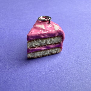 Pode incluir: Um amuleto de bolo rosa e branco com um anel prateado para pendurar. O bolo tem três camadas e cobertura rosa com granulado branco.