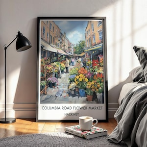 May include: A framed print of the Columbia Road Flower Market in Hackney, featuring a vibrant street scene with colorful flowers and market stalls. The print is displayed on a wooden floor, with a black lamp and a bed in the background.