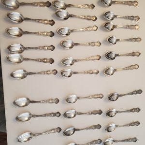 May include: A collection of 24 silver spoons with ornate designs. The spoons are arranged in rows on a white surface.