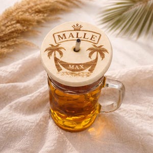 Könnte beinhalten: Ein klares Glas mit goldener Flüssigkeit, bedeckt mit einem Holzdeckel. Der Deckel zeigt die Wörter "MALLE" und "MAX" mit Palmen- und Kronenmotiven. Ein silberner Strohhalm ragt aus dem Deckel.