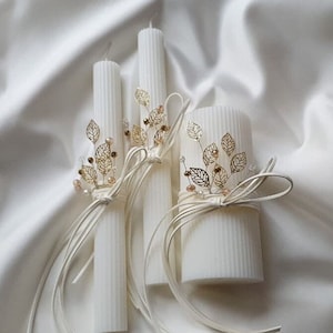 May include: Three white pillar candles with gold leaf accents and white ribbon bows. The candles are decorated with gold leaf sprigs and small white beads.