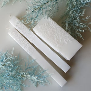 Puede incluir: Tres velas blancas en forma de pilar con un diseño floral en relieve. Las velas están dispuestas en forma escalonada sobre una superficie blanca.