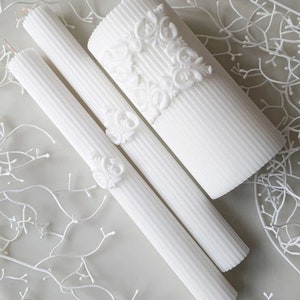 May include: Three white pillar candles with intricate raised floral designs. The candles are arranged in a row, with the largest candle in the back and the smallest candle in the front.