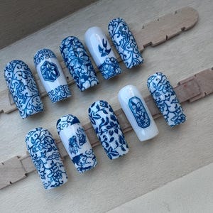 May include: A set of ten artificial nails with a blue and white floral design. The nails are arranged on two wooden sticks. The nails feature various patterns, including floral motifs and geometric shapes, reminiscent of porcelain.