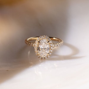 May include: A gold engagement ring featuring an oval-shaped diamond centre stone with a halo of smaller diamonds. The band is also set with smaller diamonds.