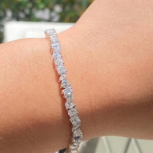 Puede incluir: Una pulsera de plata con una variedad de piedras preciosas claras y brillantes. La pulsera presenta una mezcla de formas, incluyendo piedras redondas, baguette y en forma de pera, engastadas en un diseño delicado y enlazado. La pulsera se muestra en una muñeca.