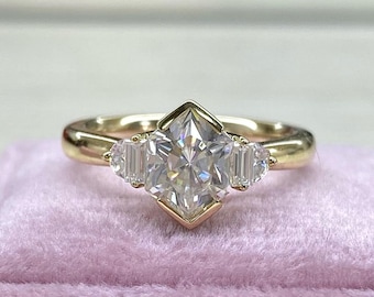 Puede incluir: Un anillo de compromiso de oro con una piedra central grande, cuadrada y transparente, y dos piedras más pequeñas de forma baguette a cada lado.