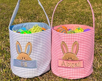 Puede incluir: Dos cestas de Pascua de tela de vichy, una azul y otra rosa, con orejas de conejo y nombres personalizados "Eli" y "Abigail".