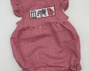 May include: A red and white gingham bubble romper featuring ruffled sleeves and a smocked chest panel. The center panel displays embroidered icons including a black arch, a grey bulldog, and a red silhouette of the state of Georgia.