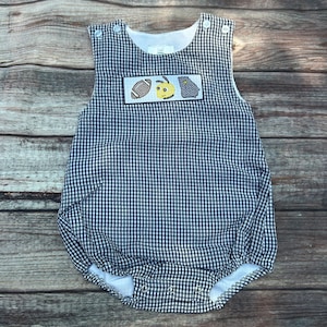 May include: A black and white gingham baby onesie with a patch on the front featuring a football, a bee, and a state of Georgia outline. The onesie has a white trim around the neck and armholes.