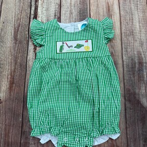 May include: Green and white gingham baby romper with ruffle sleeves and a white bottom. The romper features an embroidered golf-themed design on the chest, including a golf club, golf ball, and flag. The garment has snap closures.
