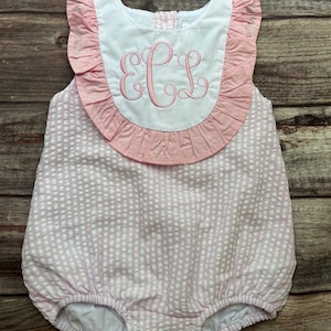 May include: A pink and white striped baby romper with a white bib-style collar. The collar is trimmed with pink ruffles and features embroidered pink initials. The romper has a bubble silhouette and snap closures.