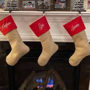 May include: Three Christmas stockings hanging above a fireplace. Each stocking is tan with a red cuff. The cuffs are embroidered with the names "Meghan", "Gus", and "Justin" in white script. The stockings are suspended from black metal hooks.