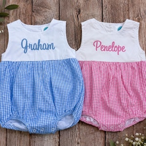 May include: Two sleeveless bubble rompers with white bodices and gingham bottoms. One romper features blue gingham with the embroidered name Graham in blue, and the other features pink gingham with the embroidered name Penelope in pink.