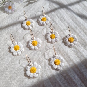 Pode incluir: Oito pares de brincos de argola dourados com pingentes de flores de margarida brancas. As margaridas t&ecirc;m centros amarelos.