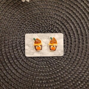 May include: A pair of brown and orange pumpkin spice latte earrings. The earrings are made of polymer clay and are shaped like a cup of coffee with a pumpkin spice topping.