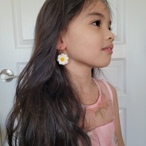 Pode incluir: Uma pessoa jovem com cabelo comprido e escuro vestindo um vestido rosa floral e brincos de margarida branca.
