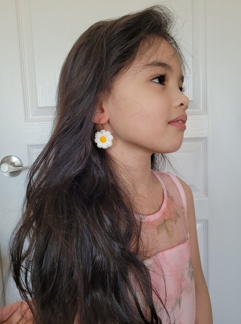 Pode incluir: Uma pessoa jovem com cabelo comprido e escuro vestindo um vestido rosa floral e brincos de margarida branca.