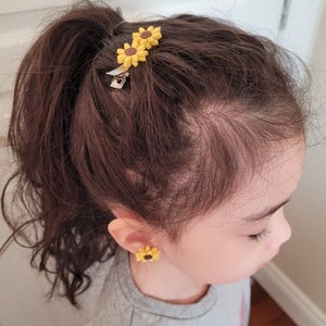 May include: A young girl with brown hair wearing a grey sweatshirt and sunflower earrings. Her hair is pulled back in a ponytail with two sunflower hair clips.