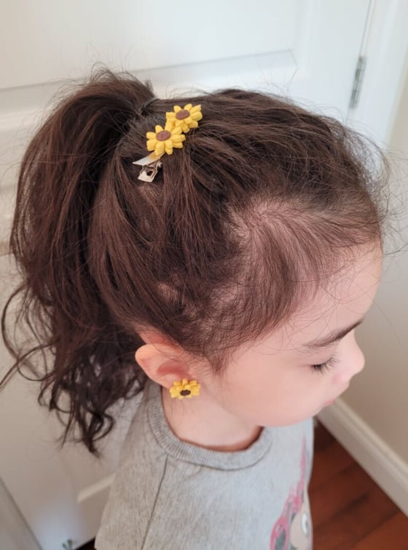 May include: A young girl with brown hair wearing a grey sweatshirt and sunflower earrings. Her hair is pulled back in a ponytail with two sunflower hair clips.