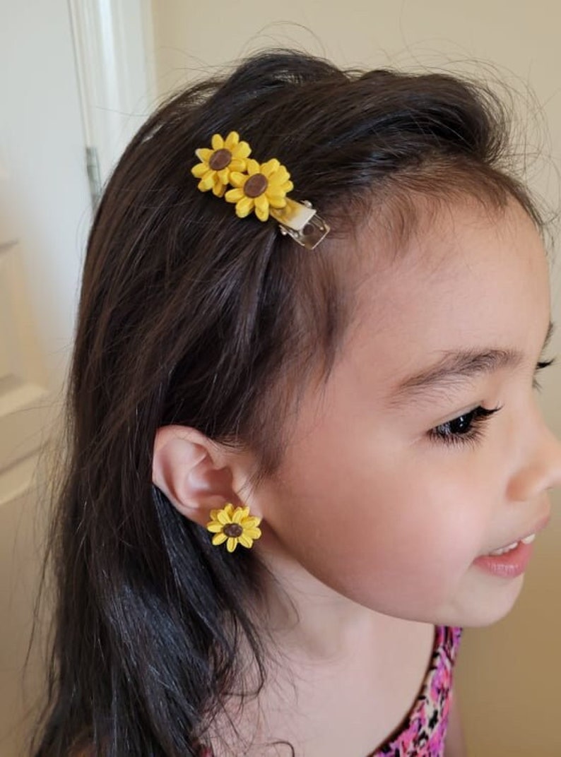 May include: A young girl wearing a pair of yellow sunflower earrings and a sunflower hair clip with two flowers.