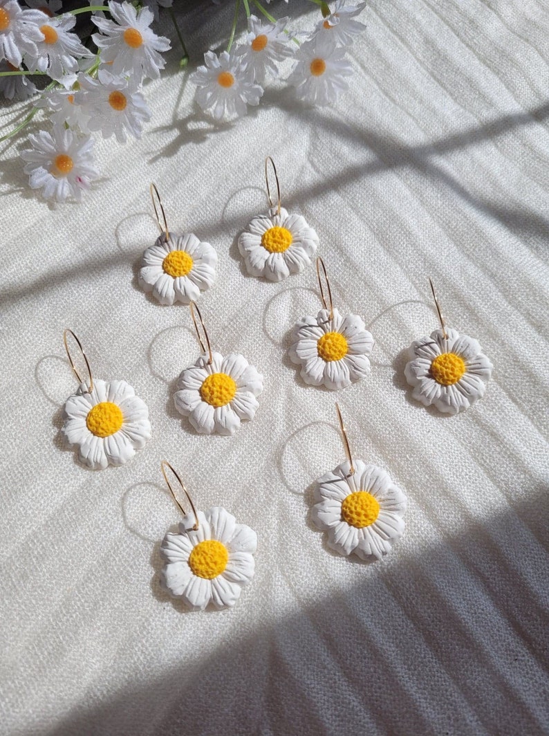 Pode incluir: Oito pares de brincos de argola dourados com pingentes de flores de margarida brancas. As margaridas t&ecirc;m centros amarelos.