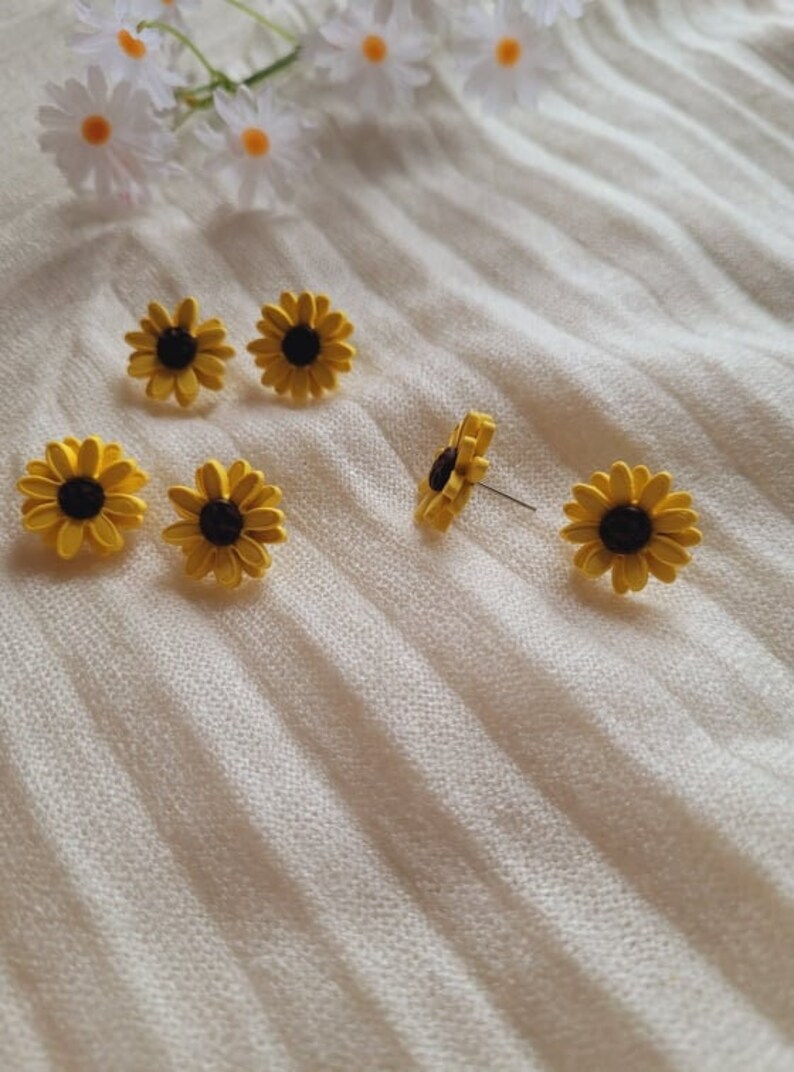 May include: Six yellow sunflower earrings with black centers. The earrings are on a white textured surface.