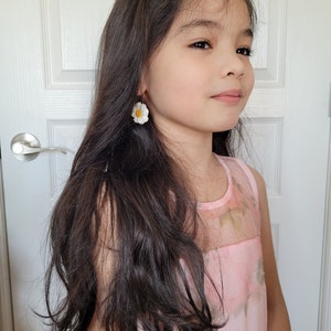Pode incluir: Uma menina jovem com cabelo comprido e escuro vestindo um vestido rosa floral e brincos de margarida branca.