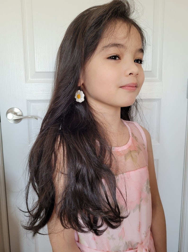 Pode incluir: Uma menina jovem com cabelo comprido e escuro vestindo um vestido rosa floral e brincos de margarida branca.