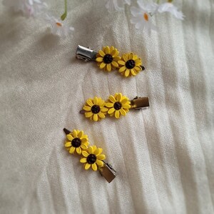 May include: Three yellow sunflower hair clips with brown centers. The clips are on a white textured surface.