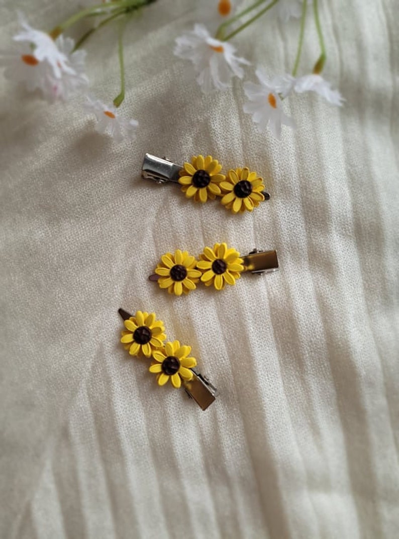 May include: Three yellow sunflower hair clips with brown centers. The clips are on a white textured surface.