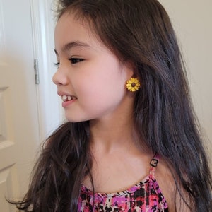 May include: A young girl wearing a pink and black patterned tank top with sunflower earrings.