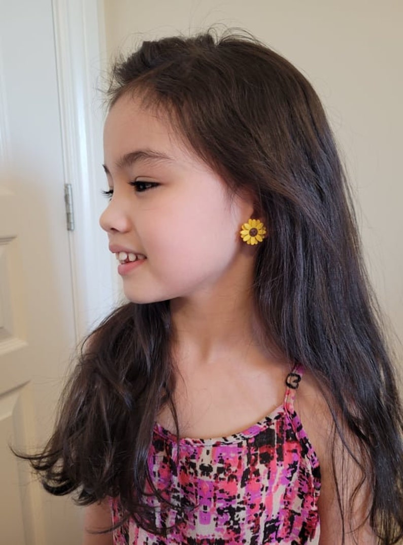 May include: A young girl wearing a pink and black patterned tank top with sunflower earrings.
