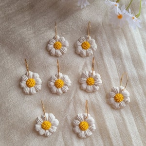 Pode incluir: Oito pares de brincos de argola dourados com pingentes de flores de margarida brancas. As margaridas t&ecirc;m centros amarelos.