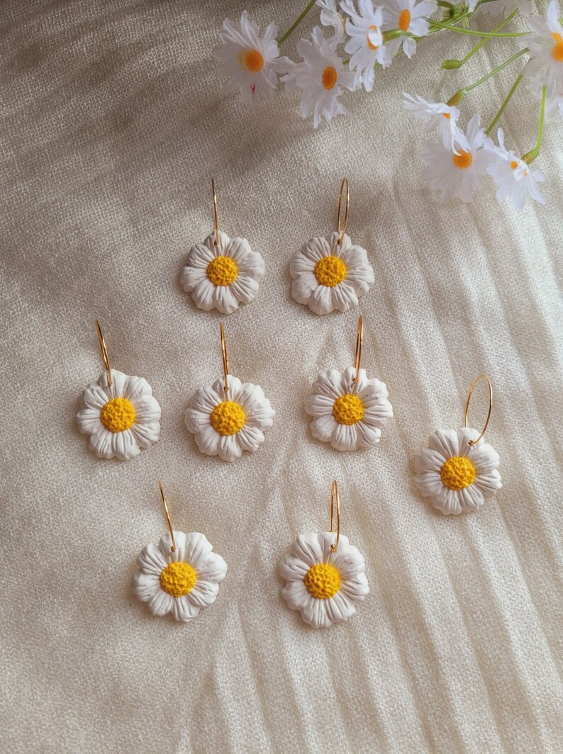 Pode incluir: Oito pares de brincos de argola dourados com pingentes de flores de margarida brancas. As margaridas t&ecirc;m centros amarelos.