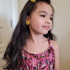 May include: A young girl with long dark hair wearing a pink and black patterned sleeveless dress. She has a yellow sunflower hair clip in her hair and yellow sunflower earrings.