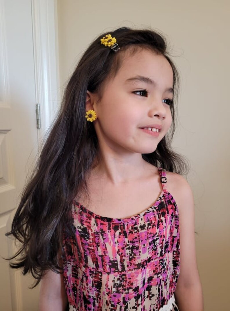 May include: A young girl with long dark hair wearing a pink and black patterned sleeveless dress. She has a yellow sunflower hair clip in her hair and yellow sunflower earrings.