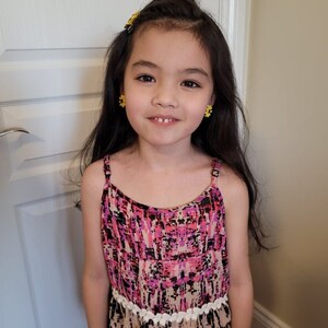 May include: A young girl with long dark hair wearing a pink and black patterned sleeveless dress with a white crocheted trim. She is smiling and has yellow flower-shaped earrings.
