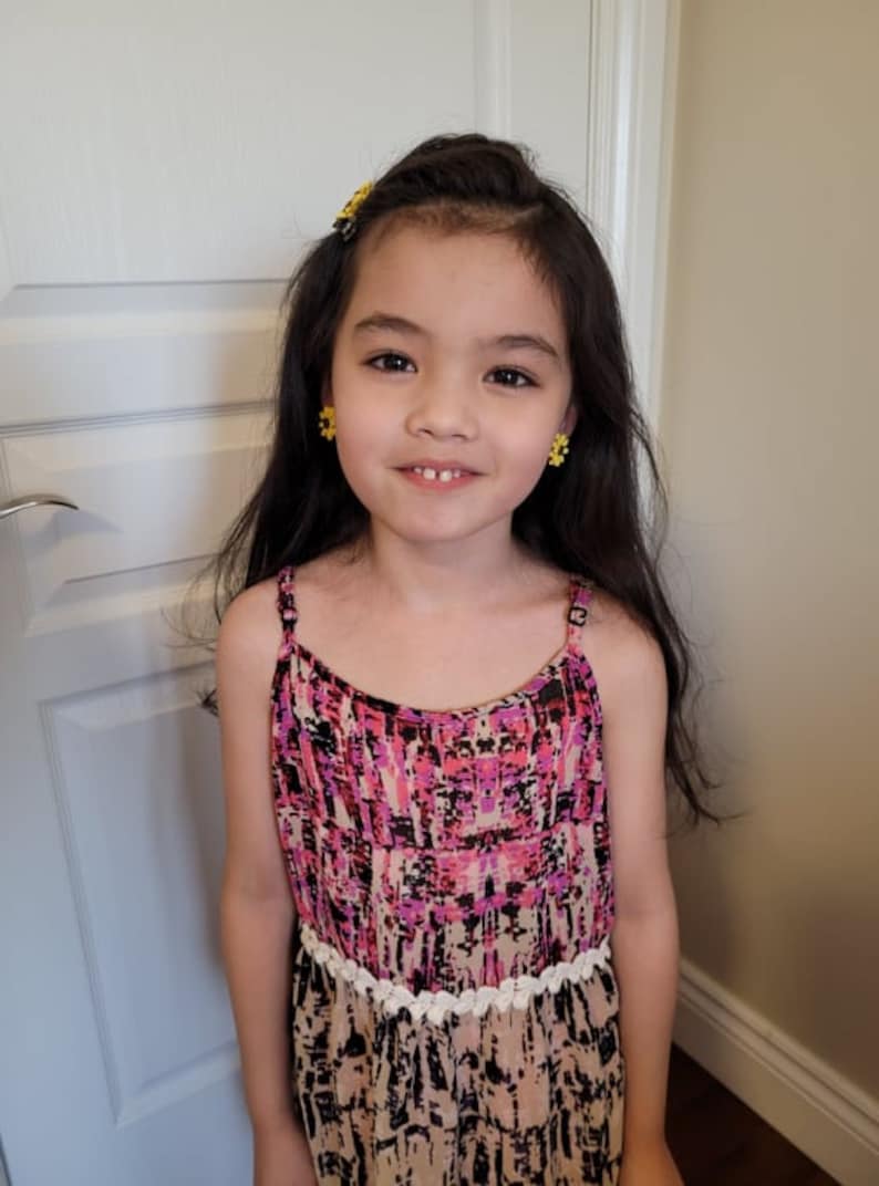 May include: A young girl with long dark hair wearing a pink and black patterned sleeveless dress with a white crocheted trim. She is smiling and has yellow flower-shaped earrings.