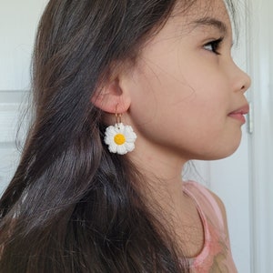 Pode incluir: Uma pessoa jovem usando um par de brincos de argola dourados com pingentes de flores de margarida brancas. A margarida tem um centro amarelo.