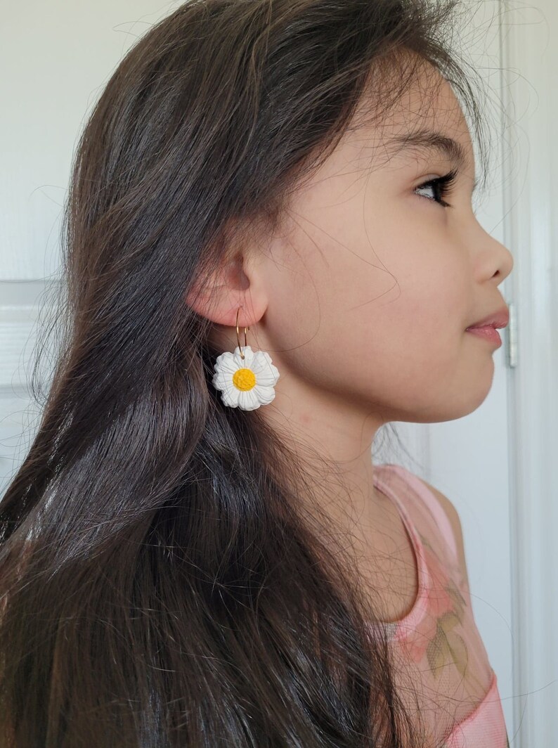 Pode incluir: Uma pessoa jovem usando um par de brincos de argola dourados com pingentes de flores de margarida brancas. A margarida tem um centro amarelo.