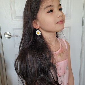 Pode incluir: Uma menina jovem com cabelo comprido e escuro vestindo um vestido rosa floral e brincos de margarida branca.