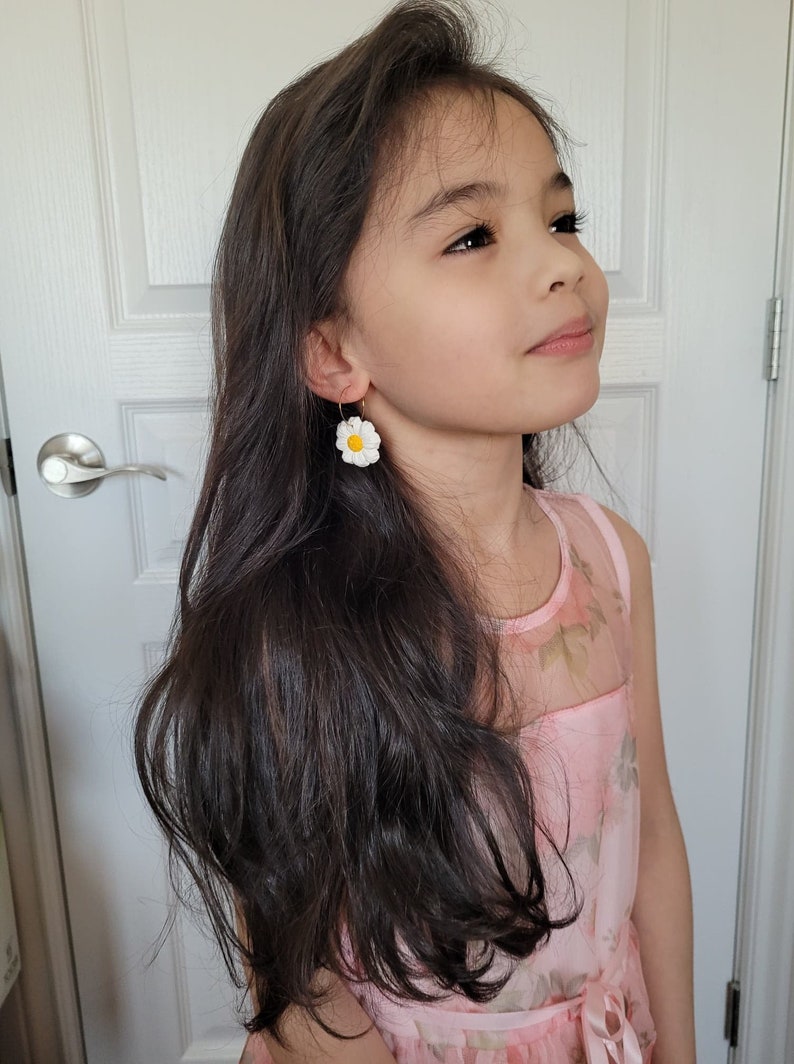 Pode incluir: Uma menina jovem com cabelo comprido e escuro vestindo um vestido rosa floral e brincos de margarida branca.