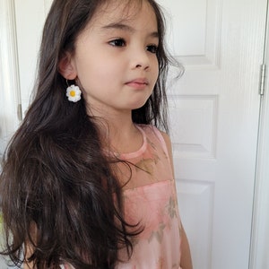 Pode incluir: Uma menina jovem com cabelo comprido e escuro vestindo um vestido rosa floral e brincos de margarida branca.
