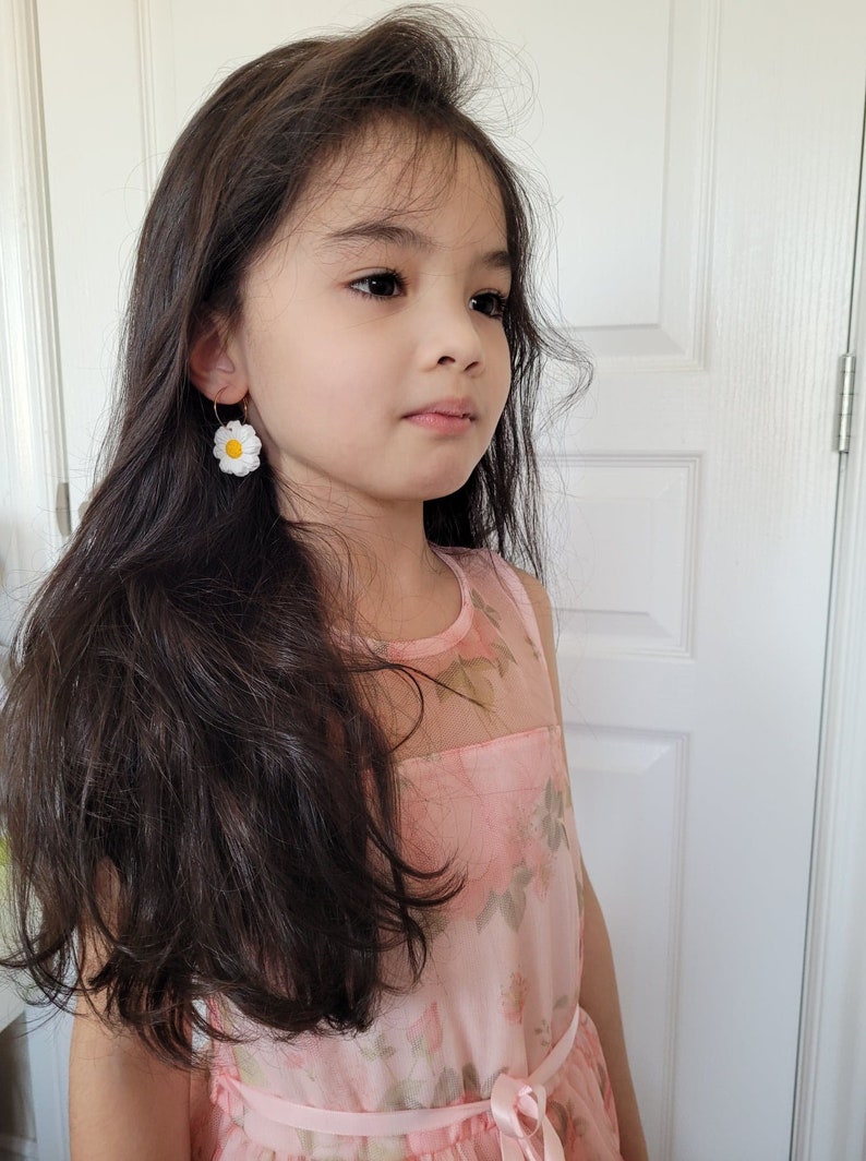 Pode incluir: Uma menina jovem com cabelo comprido e escuro vestindo um vestido rosa floral e brincos de margarida branca.