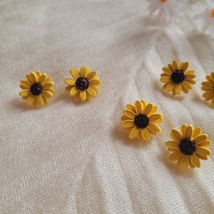 May include: Six yellow sunflower earrings with brown centers. The earrings are on a white fabric background.