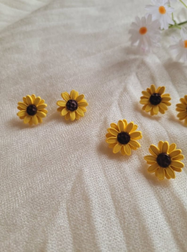 May include: Six yellow sunflower earrings with brown centers. The earrings are on a white fabric background.